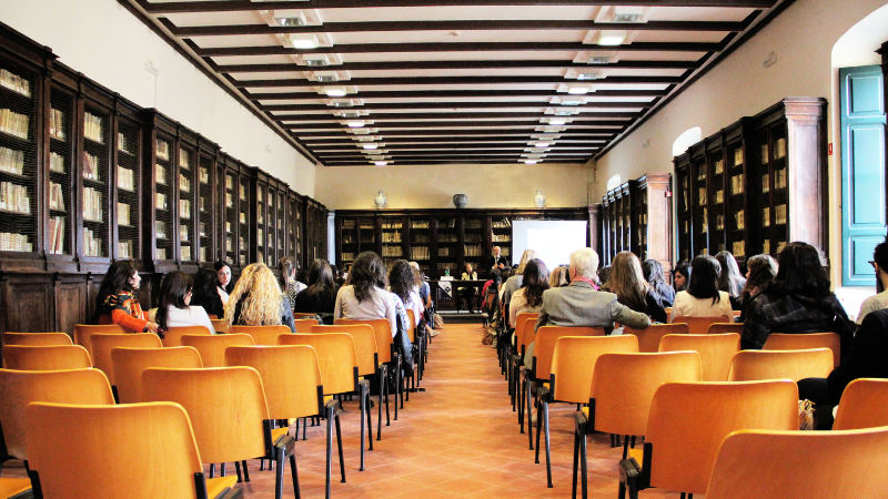 Een groep mensen zittend op oranje stoelen tijdens een presentatie in een zaal met boekenkasten langs de muren.